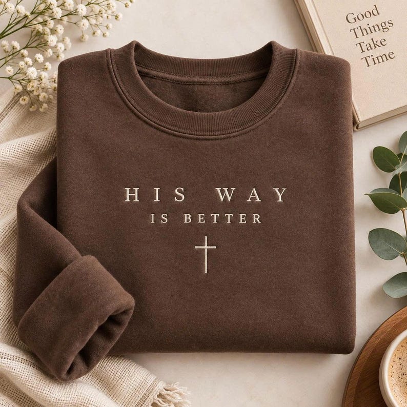 May include: A dark brown crewneck sweatshirt featuring the embroidered text "HIS WAY IS BETTER" in a light cream color, positioned above a small, simple cross symbol centered on the chest of the garment.