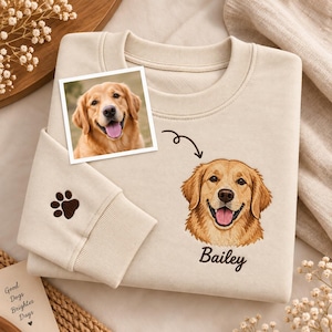 May include: A beige crewneck sweatshirt featuring an embroidered golden retriever portrait with the name Bailey underneath. A small brown paw print is embroidered on the sleeve cuff. A square photograph of the same dog is placed on the fabric.
