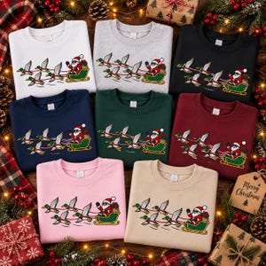 May include: A collection of eight crewneck sweatshirts in white, gray, black, navy, dark green, maroon, pink, and beige. Each sweatshirt features a graphic of Santa Claus in a green sleigh pulled by a team of flying mallard ducks in flight.