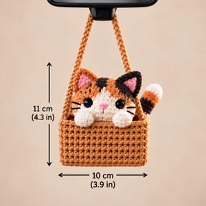 May include: A crocheted calico cat figurine sitting inside a brown rectangular basket with a hanging strap. The cat has black, orange, and white fur, pink ears, and black eyes. The basket measures 3.9 inches wide and 4.3 inches in total height.