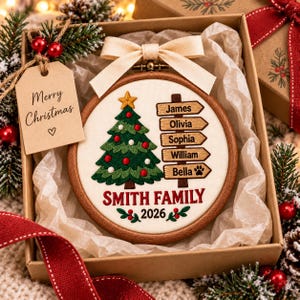 May include: A round wooden embroidery hoop ornament featuring a stitched green Christmas tree with red and white ornaments, a yellow star, and a signpost with the names James, Olivia, Sophia, William, and Bella. Text reads Smith Family 2026.