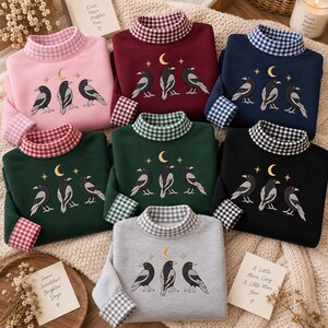 May include: A collection of seven long-sleeve sweatshirts in pink, maroon, navy, dark green, black, and gray. Each features a mock neck with a gingham pattern, matching cuffs, and an embroidered design of three crows under a crescent moon.