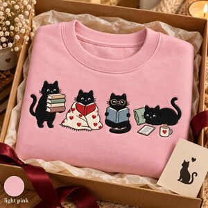 May include: A light pink crewneck sweatshirt featuring an embroidered graphic of four black cats. One cat holds a stack of books, one is wrapped in a heart-patterned blanket reading, one wears glasses while reading, and one lies by a pillow and mug.