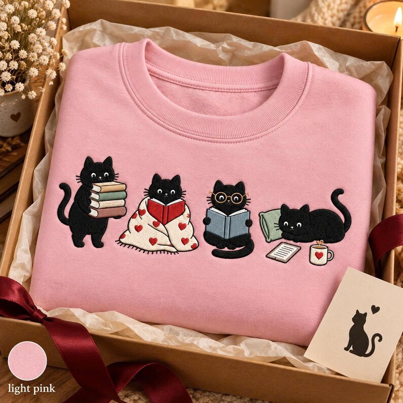 May include: A light pink crewneck sweatshirt featuring an embroidered graphic of four black cats. One cat holds a stack of books, one is wrapped in a heart-patterned blanket reading, one wears glasses while reading, and one lies by a pillow and mug.