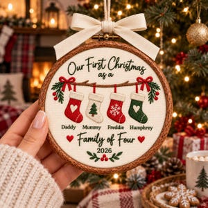 May include: A round wooden embroidery hoop ornament featuring cream fabric with embroidered text Our First Christmas as a Family of Four 2026. It displays four hanging stockings labeled Daddy, Mummy, Freddie, and Humphrey with a cream bow.