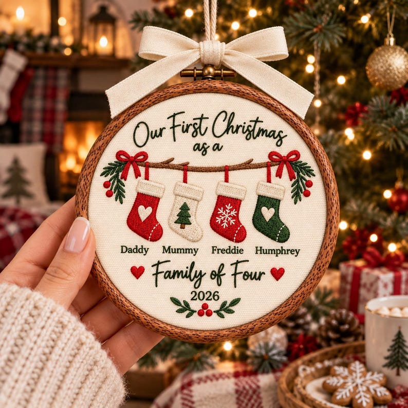 May include: A round wooden embroidery hoop ornament featuring cream fabric with embroidered text Our First Christmas as a Family of Four 2026. It displays four hanging stockings labeled Daddy, Mummy, Freddie, and Humphrey with a cream bow.