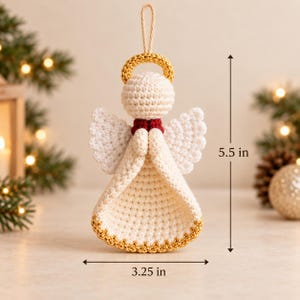 May include: A crocheted angel ornament featuring a cream-colored body, white wings, a gold halo, and a red collar. The figure has a loop for hanging and measures 5.5 inches in height and 3.25 inches in width as indicated by the dimension markers.