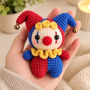May include: A small crochet clown doll featuring a red and blue jester hat with two small metal bells at the tips. The doll has a cream-colored face, a red nose, black eyes, blue tear-drop markings, yellow hair curls, and a yellow ruffled collar.