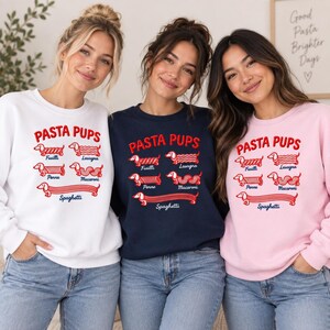 May include: Three crewneck sweatshirts in white, navy blue, and light pink. Each features red embroidered text reading PASTA PUPS above illustrations of dachshunds shaped like pasta types labeled Fusilli, Lasagna, Penne, Macaroni, and Spaghetti.
