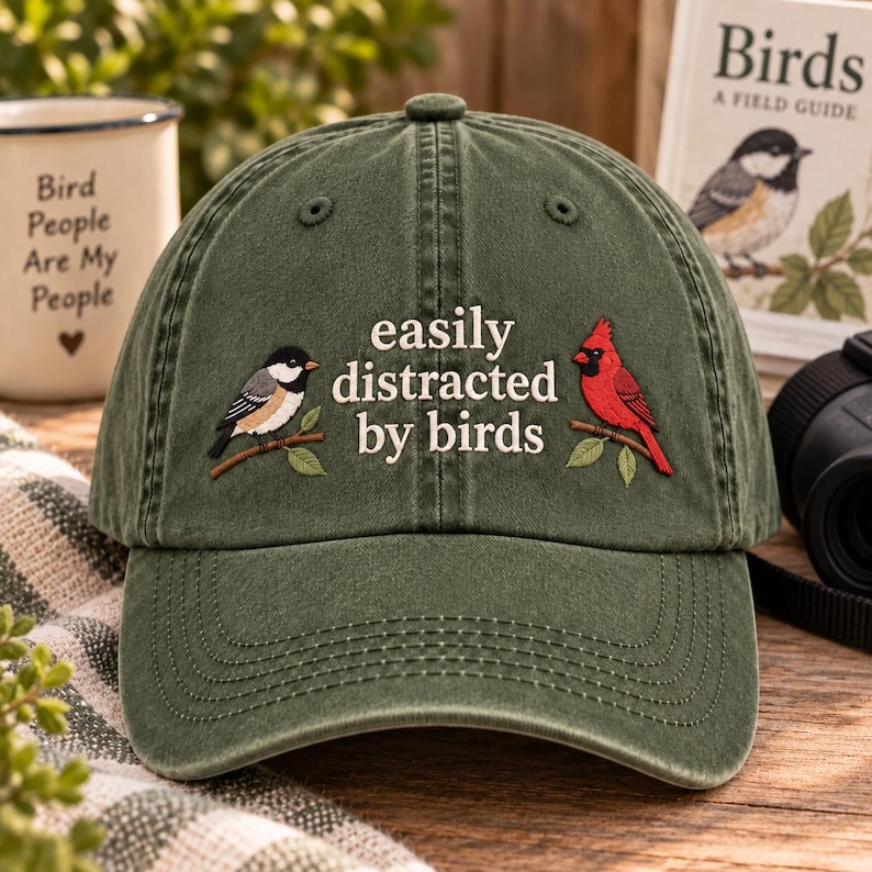 May include: An olive green baseball cap featuring embroidered details of a black-capped chickadee on the left and a bright red cardinal on the right, with the text "easily distracted by birds" stitched in white lettering across the center front.