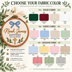 May include: A circular wooden embroidery hoop featuring a beige fabric with a blue bow, the name Noah James, the word BAPTIZED, a date, and a cross. A color chart displays 15 fabric options including solid, striped, and gingham patterns.
