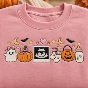May include: A dusty pink crewneck sweatshirt featuring an embroidered design of a ghost with a bow, a pumpkin with a bow, a sonogram image, baby socks, a jack-o'-lantern bucket, and a baby bottle, accented with stars, moons, bats, and hearts.