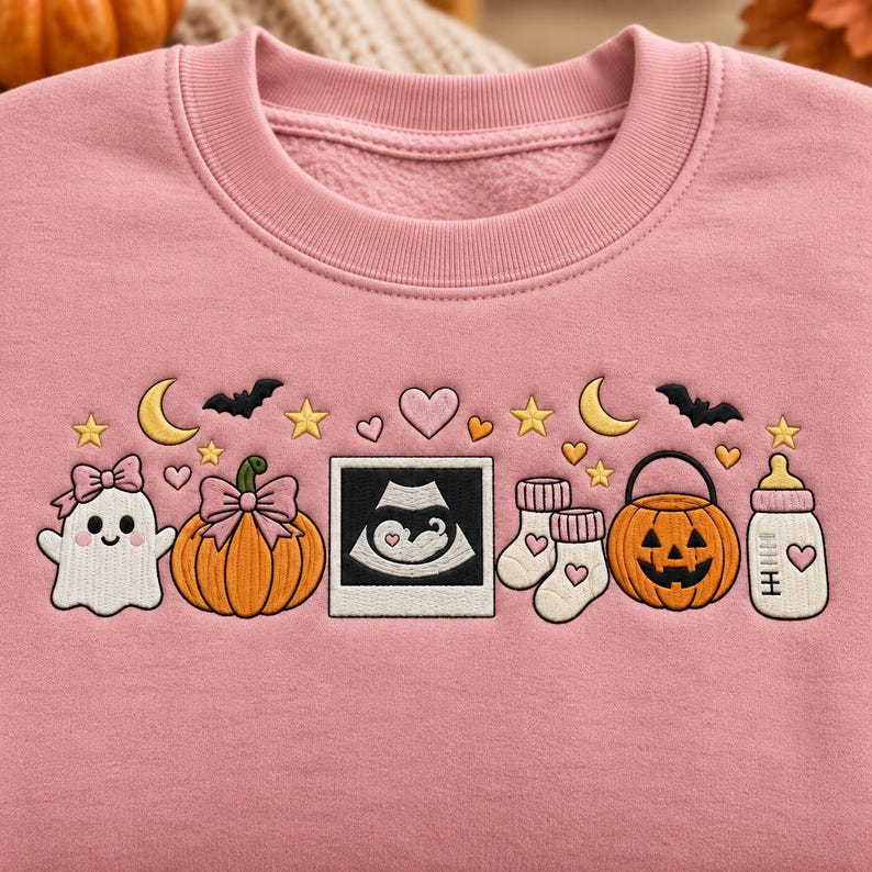 May include: A dusty pink crewneck sweatshirt featuring an embroidered design of a ghost with a bow, a pumpkin with a bow, a sonogram image, baby socks, a jack-o'-lantern bucket, and a baby bottle, accented with stars, moons, bats, and hearts.