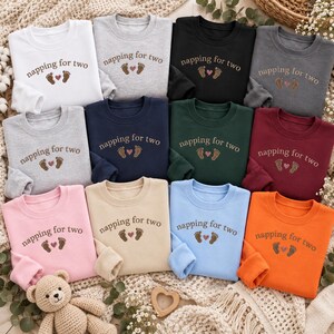May include: A collection of twelve crewneck sweatshirts in various colors including white, gray, black, navy, green, maroon, pink, beige, light blue, and orange. Each sweatshirt features the embroidered text napping for two with two baby footprints and a small heart.