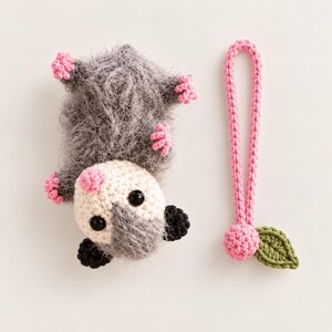 May include: A crocheted gray fuzzy opossum plush toy with a cream-colored face, black eyes, and pink paws, accompanied by a separate pink crocheted loop strap featuring a small pink sphere and a single green crocheted leaf attached to the end.