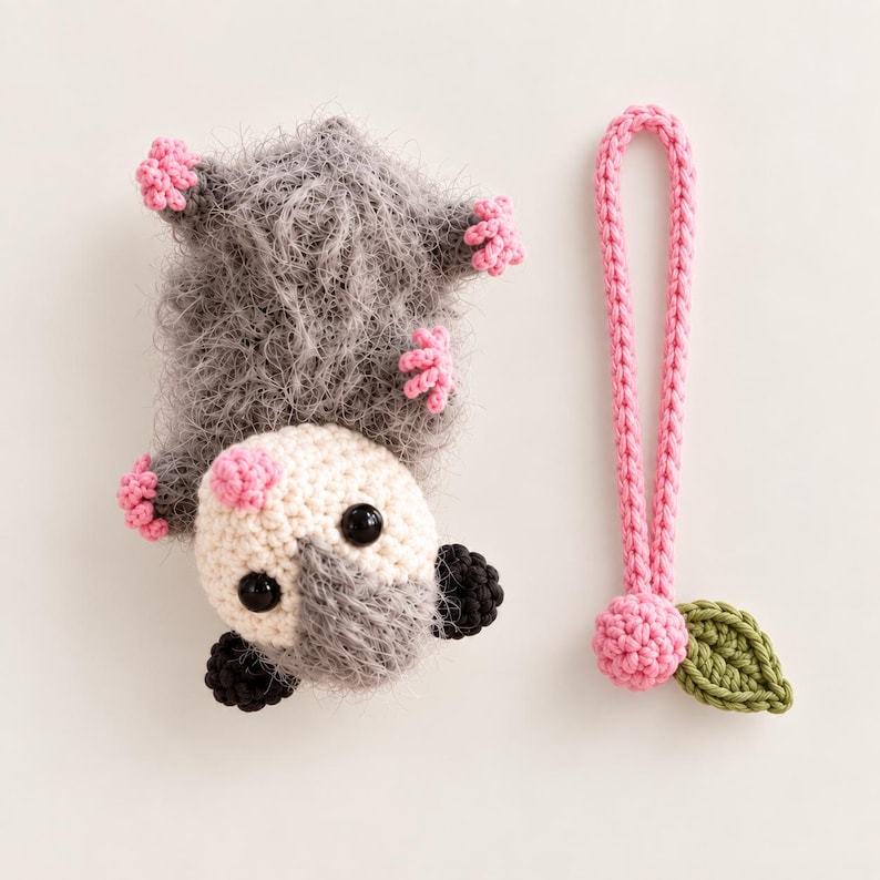 May include: A crocheted gray fuzzy opossum plush toy with a cream-colored face, black eyes, and pink paws, accompanied by a separate pink crocheted loop strap featuring a small pink sphere and a single green crocheted leaf attached to the end.