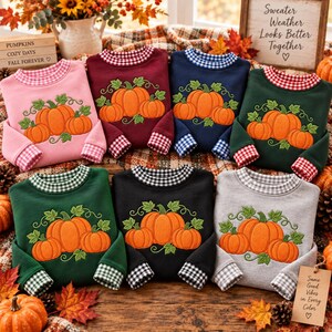 May include: A collection of seven long-sleeve sweatshirts in pink, maroon, navy blue, dark green, forest green, black, and gray. Each sweatshirt features a central embroidered pumpkin patch design and a gingham-patterned collar and sleeve cuffs.