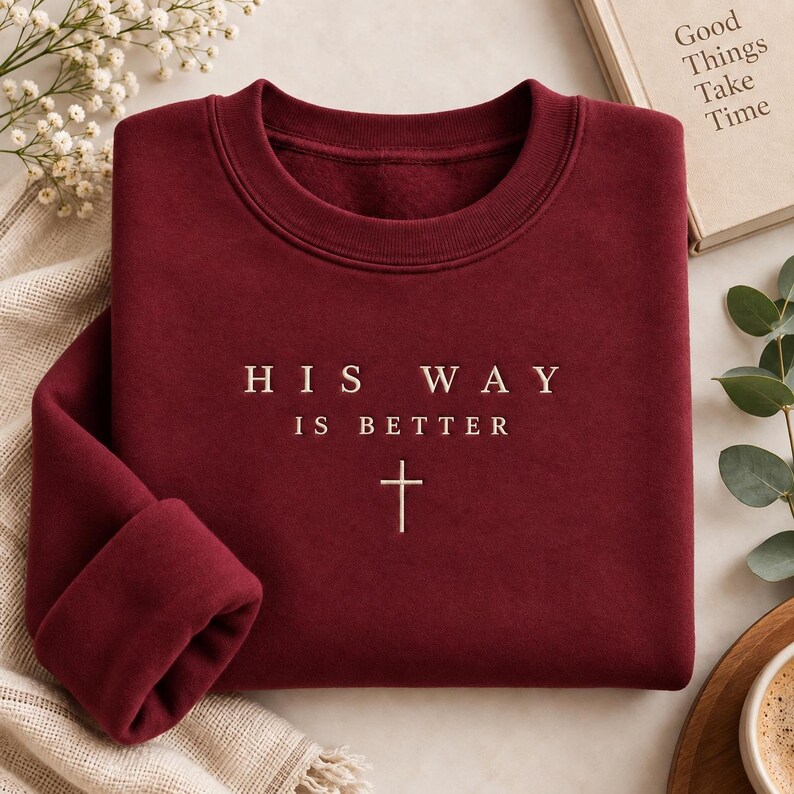 May include: A deep maroon crewneck sweatshirt featuring the embroidered text HIS WAY IS BETTER in a light cream color, positioned above a small, simple cross symbol. The garment has a ribbed collar and long sleeves with ribbed cuffs.