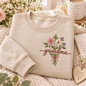 May include: A beige crewneck sweatshirt featuring an embroidered bouquet of pink, white, and purple flowers tied with a pink ribbon. The word grandma is stitched in brown cursive script below the flowers. The fabric has a soft, fleece-like texture.