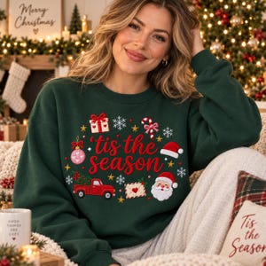 May include: A dark green long-sleeve sweatshirt featuring the red embroidered text 'tis the season' surrounded by festive icons including a gift box, candy cane, pink ornament, red truck carrying a Christmas tree, envelope, and Santa Claus face.