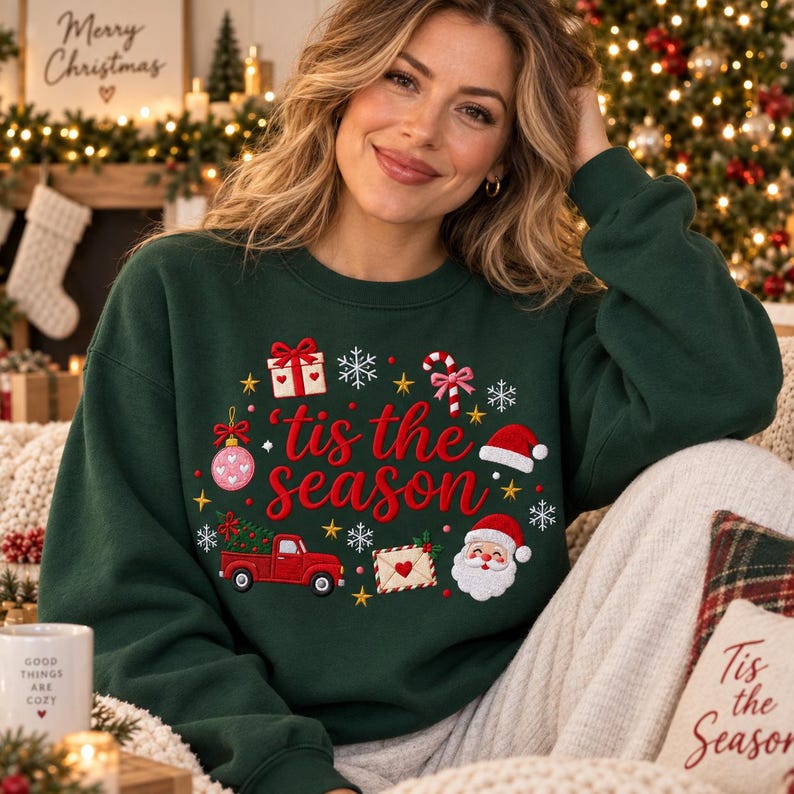 May include: A dark green long-sleeve sweatshirt featuring the red embroidered text 'tis the season' surrounded by festive icons including a gift box, candy cane, pink ornament, red truck carrying a Christmas tree, envelope, and Santa Claus face.