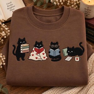 May include: A brown crewneck sweatshirt featuring an embroidered graphic of four black cats. One cat holds a stack of books, one is wrapped in a heart-patterned blanket reading, one wears round glasses reading a book, and one lies by a mug.