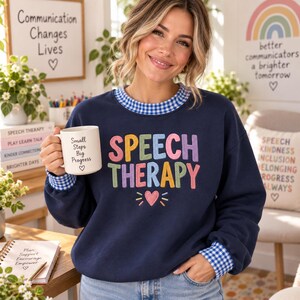 May include: A navy blue crewneck sweatshirt with a blue and white gingham collar and cuffs. The front features the text "SPEECH THERAPY" in colorful, embroidered letters above a small pink heart. A white mug displays "Small Steps Big Progress".