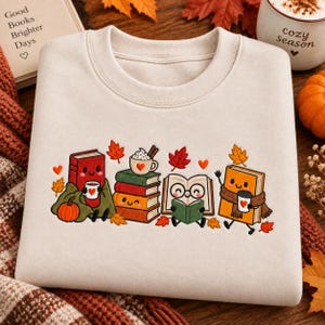 May include: A cream-colored crewneck sweatshirt featuring a colorful embroidered graphic of anthropomorphic books. The design includes books with faces, a stack of books topped with a mug, autumn leaves, and small hearts on a light fabric.