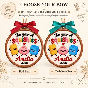 May include: Two circular wooden embroidery hoops featuring embroidered text The year of squishies and Amelia 2026 with three cartoon characters. One hoop has a red satin bow and the other has a teal green satin bow, both with hanging twine.