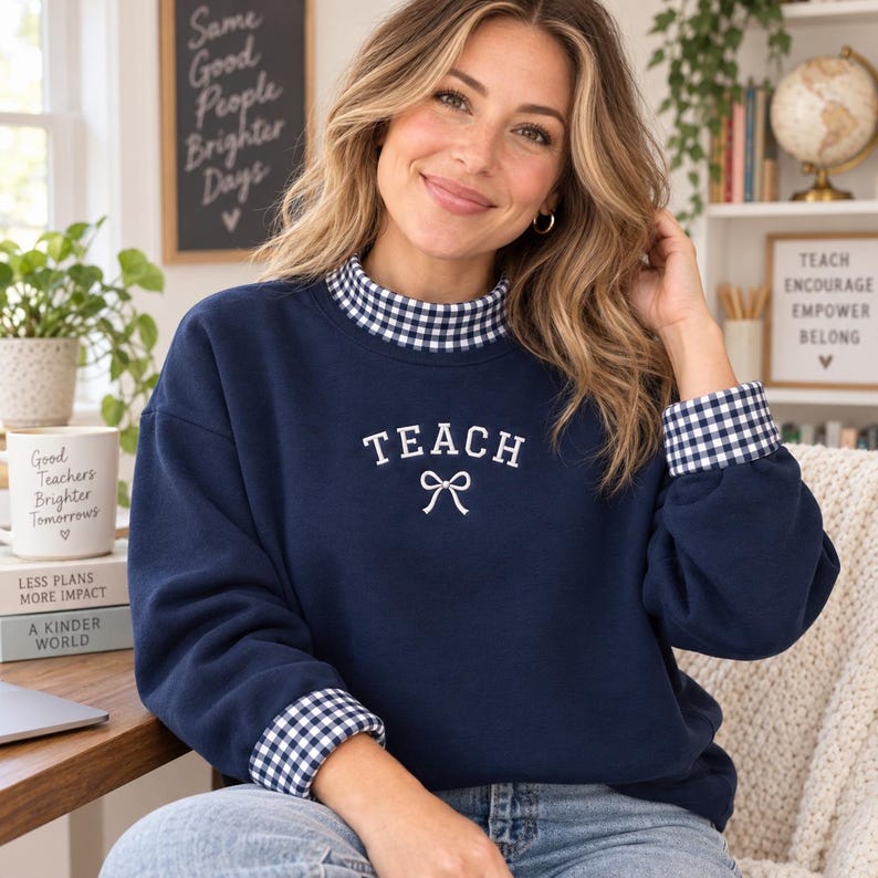 May include: A navy blue long-sleeve sweatshirt featuring a white embroidered text that reads TEACH with a small bow icon underneath. The garment has a mock neck collar and sleeve cuffs in a navy and white gingham check pattern.