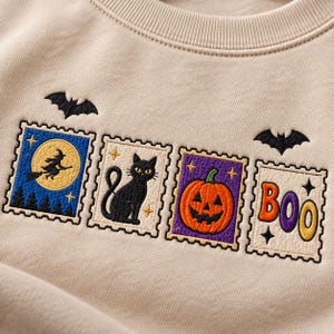May include: A beige sweatshirt featuring embroidered designs of four rectangular postage stamps. The stamps depict a witch on a broomstick, a black cat, a jack-o'-lantern, and the word BOO in colorful letters, accented by two black bats.