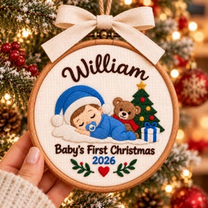 May include: A round wooden embroidery hoop ornament featuring a cream fabric background with a beige ribbon bow at the top. The embroidery depicts a sleeping baby in a blue outfit and hat, a brown teddy bear, a Christmas tree, and a gift box. Text reads William, Baby's First Christmas, and 2026, with green leaves and a red heart.