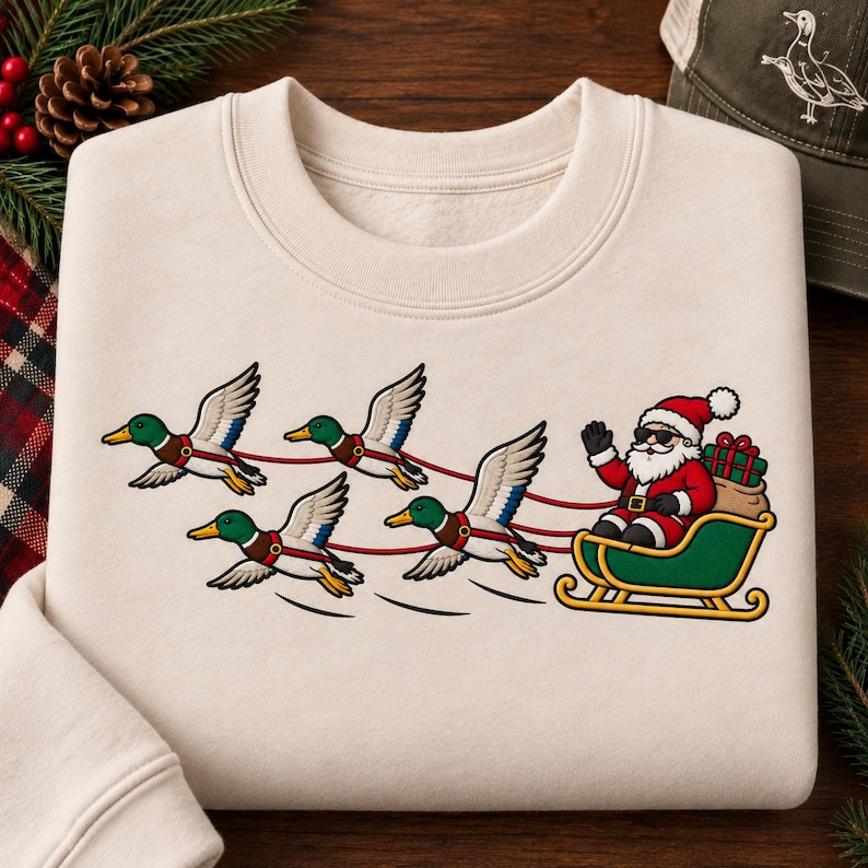May include: A cream-colored crewneck sweatshirt featuring a graphic print of Santa Claus wearing sunglasses and waving while sitting in a green sleigh. The sleigh is being pulled by four mallard ducks with red harnesses, and a sack of gifts is in the back.