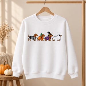 May include: A white long-sleeve crewneck sweatshirt featuring a graphic print of four brown dachshunds in Halloween costumes. The dogs are dressed as a skeleton, a pumpkin, a witch with a purple cape and hat, and a ghost under a white sheet.