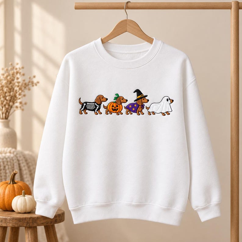 May include: A white long-sleeve crewneck sweatshirt featuring a graphic print of four brown dachshunds in Halloween costumes. The dogs are dressed as a skeleton, a pumpkin, a witch with a purple cape and hat, and a ghost under a white sheet.