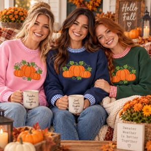 May include: Three crewneck sweatshirts in pink, navy blue, and dark green, each featuring an embroidered pumpkin graphic and a gingham collar and cuff trim. Each sweatshirt is paired with a ceramic mug with printed text, including Pumpkin Spice Everything, Good Friends Brighter Days, and Sweater Weather Is Better Together.