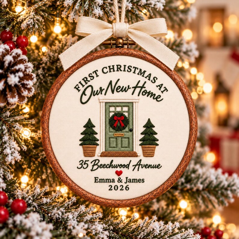 May include: A round wooden embroidery hoop ornament featuring white fabric with embroidered text First Christmas at Our New Home, 35 Beechwood Avenue, Emma & James, 2026, a green front door with a wreath, two small trees, and a cream ribbon bow.