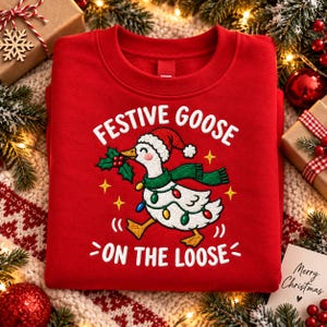 May include: A red crewneck sweatshirt featuring an embroidered graphic of a white goose wearing a Santa hat, a green scarf, and a string of colorful Christmas lights. The text reads "FESTIVE GOOSE ON THE LOOSE" in white capital letters.