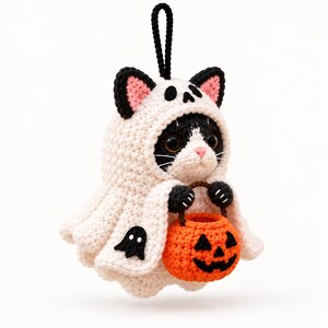 May include: A crocheted black and white cat ornament wearing a cream-colored ghost costume with a hood, featuring pink inner ears and a skull detail. The cat holds a small, round, orange crocheted jack-o'-lantern bucket with a black face.