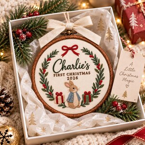 May include: A round wooden embroidery hoop featuring a cream fabric centre with a stitched wreath, a red bow, and a rabbit wearing a blue coat. The text reads Charlie's FIRST CHRISTMAS 2026. A beige gift tag reads A Little Christmas Magic with a tree icon.