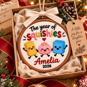 May include: A round wooden embroidery hoop ornament featuring embroidered text The year of squishies and Amelia 2026 with three colorful cartoon characters. A cream-colored ribbon bow is attached to the top, and a brown paper tag reads A little joy for a brighter year.