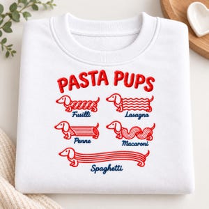 May include: A white crewneck sweatshirt featuring red embroidered graphics of dachshunds shaped like pasta varieties. The text reads PASTA PUPS above illustrations labeled Fusilli, Lasagna, Penne, Macaroni, and Spaghetti in blue cursive lettering.