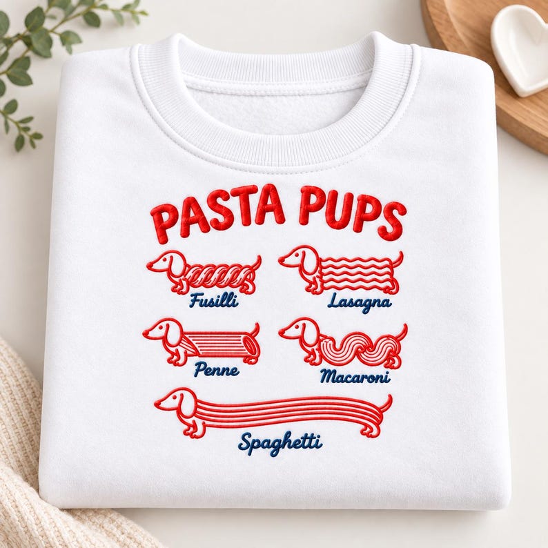 May include: A white crewneck sweatshirt featuring red embroidered graphics of dachshunds shaped like pasta varieties. The text reads PASTA PUPS above illustrations labeled Fusilli, Lasagna, Penne, Macaroni, and Spaghetti in blue cursive lettering.