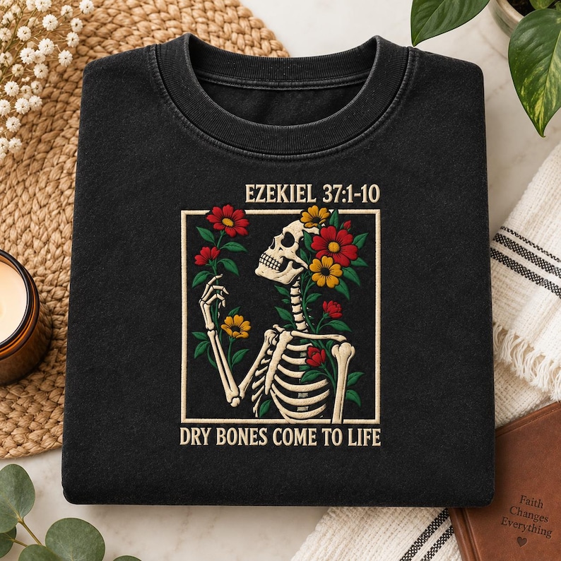 May include: A black crewneck sweatshirt featuring a graphic print of a skeleton surrounded by red and yellow flowers. The text "EZEKIEL 37:1-10" is printed above the graphic, and the text "DRY BONES COME TO LIFE" is printed below the graphic.