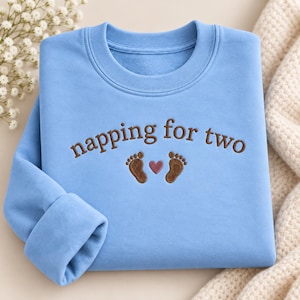 May include: A light blue crewneck sweatshirt featuring dark brown embroidered text that reads napping for two, positioned above two small brown embroidered baby footprints and a small pink heart centered between the footprints on the chest.