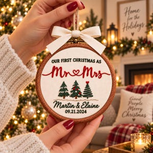 May include: A round wooden embroidery hoop ornament featuring white fabric with the embroidered text OUR FIRST CHRISTMAS AS Mr & Mrs, Martin & Elaine, and 09.21.2024. Three embroidered Christmas trees and a cream-colored fabric bow are included.