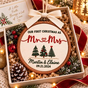 May include: A round wooden embroidery hoop ornament featuring cream fabric with embroidered text "OUR FIRST CHRISTMAS AS Mr & Mrs", "Martin & Elaine", and "09.21.2024". It includes three embroidered pine trees and a cream ribbon bow.