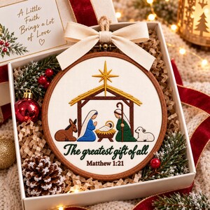 May include: A round wooden embroidery hoop featuring a nativity scene with a gold star, a manger, and figures in blue and green robes. The text reads, "The greatest gift of all" and "Matthew 1:21" in dark green and brown script on white fabric.