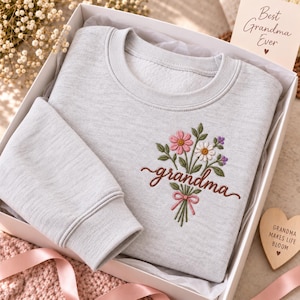 May include: A light gray crewneck sweatshirt featuring an embroidered bouquet of pink and white flowers with green stems and the word grandma in brown cursive script. The sweatshirt is inside a white gift box with a card reading Best Grandma Ever.