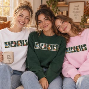 May include: Three crewneck sweatshirts in white, dark green, and light pink, each featuring a horizontal graphic of four postage stamp-style illustrations of ducks reading books. The white sweatshirt is paired with a beige mug with the text Good Books Brighter Days.